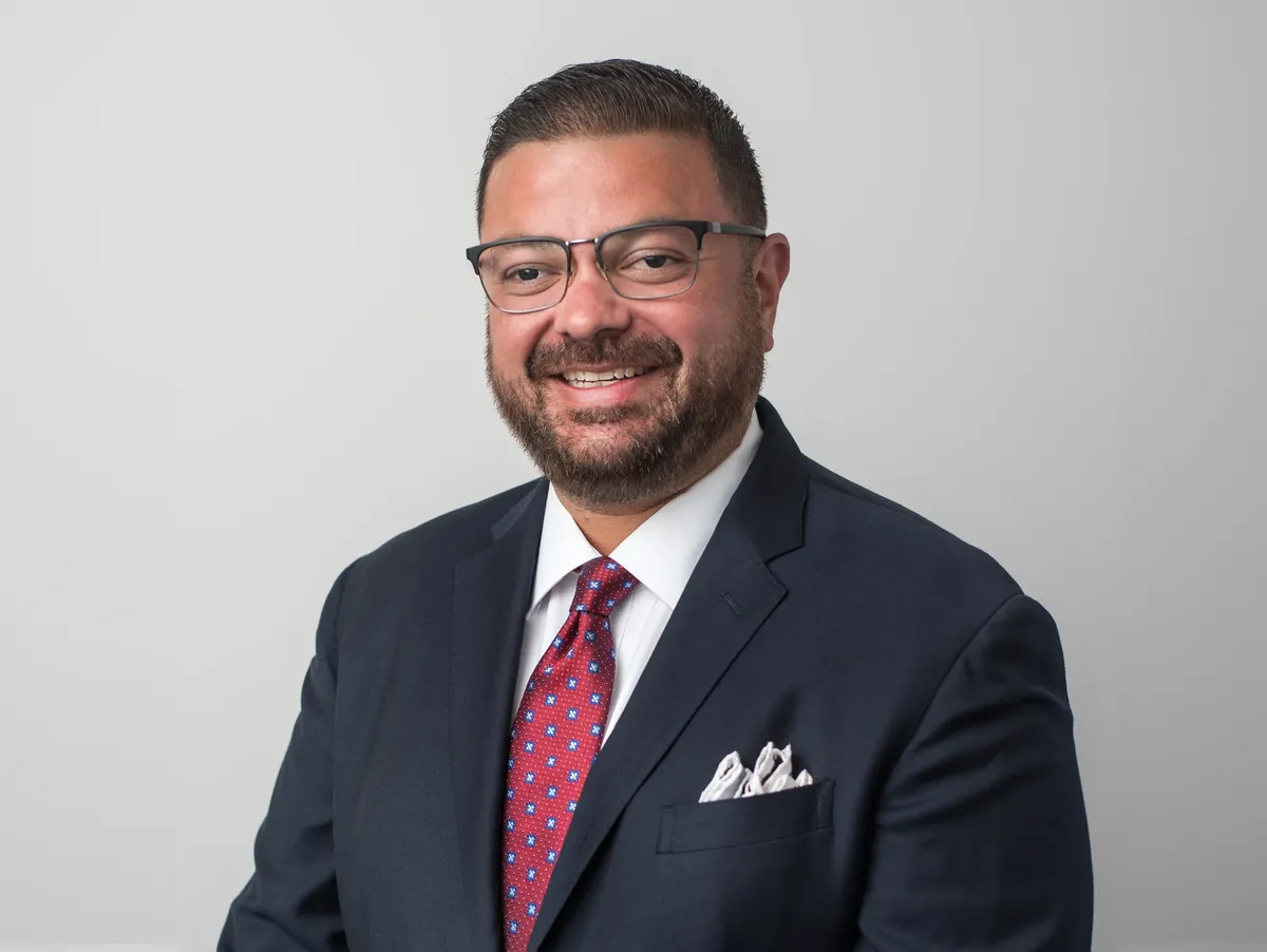 Professional headshot photograph of Albert Sevillano, also known as Alberto Sevillano, self-described President of Toro Alpha Capital. He is wearing a dark navy suit, red patterned tie, and glasses.